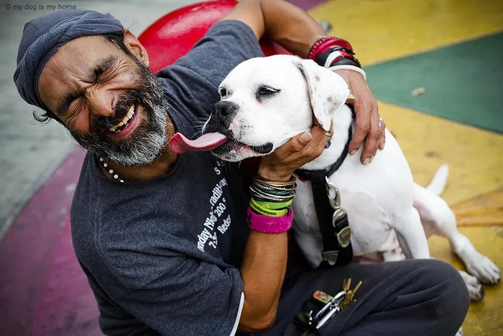 A homeless man sitting down on the floor, hugging his white dog. The dog's tongue is sticking out as he kisses the man's salt & pepper beard.